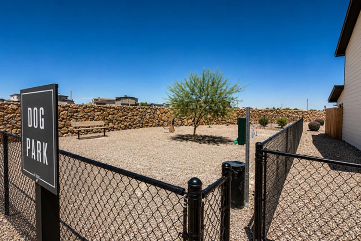 A fenced dog park featuring a gravel surface, a small tree, and benches. A sign indicating "Dog Park" stands at the entrance. In the background, there are rock walls, and the sky is clear and blue. The area is well-maintained, suitable for dogs to play and roam freely.