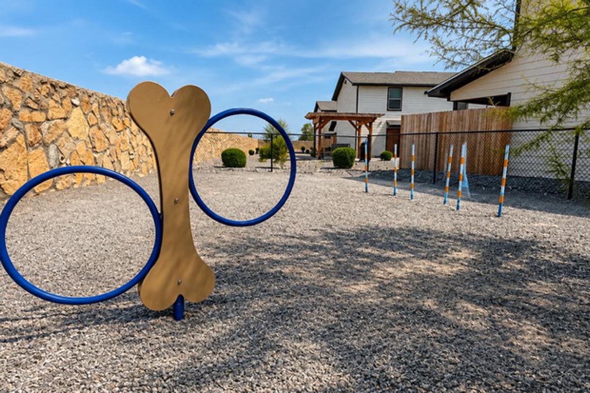 A dog agility playground featuring a large bone-shaped jump and colorful poles on a gravel surface, surrounded by a fence. In the background, a residential building can be seen under a clear blue sky with a few trees nearby.