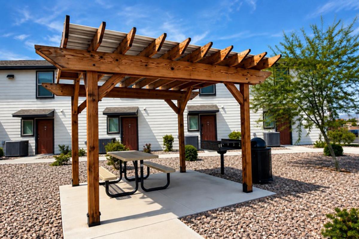 A wooden pergola with a picnic table underneath, located in a landscaped area with gravel and shrubs. In the background, there are two residential units with brown doors and air conditioning units. The sky is clear with a few clouds, creating a bright, inviting atmosphere.