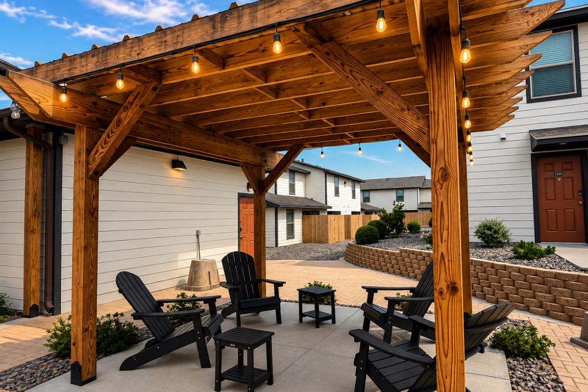 A cozy outdoor seating area featuring a wooden pergola with string lights, surrounded by neatly arranged potted plants and flower beds. Four black Adirondack chairs and a small table are set on a paved surface, with nearby buildings visible in the background. The scene is inviting and well-maintained.
