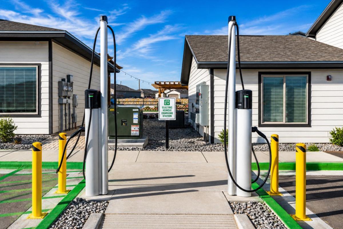 Two electric vehicle charging stations are positioned at the entrance of a building. The charging units have cables hanging down, and the area is marked with green and yellow painted lines on the ground. A sign is visible indicating it’s an electric vehicle charging station, set against a clear blue sky.