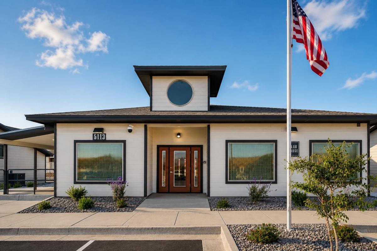 A modern single-story building with a flat roof design, featuring large windows and a circular window on the front. The entrance has double doors, and the building is surrounded by neatly landscaped gravel and plants. An American flag is positioned in front of the building.