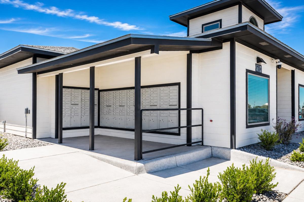 A modern building with a flat roof and white exterior. In front, there is a covered area with multiple mailboxes arranged neatly. The space features a wheelchair-accessible ramp and is surrounded by low shrubs and a small concrete walkway. Blue skies and a few clouds are visible above.