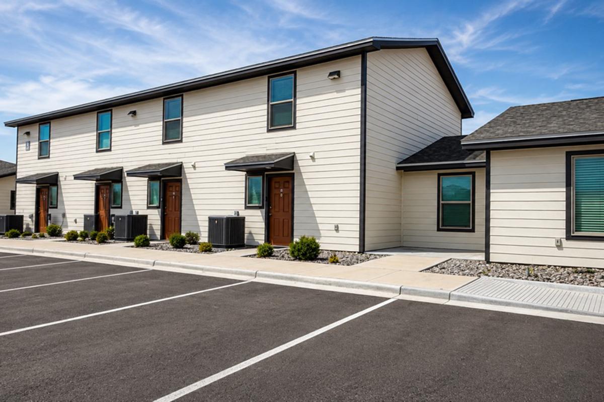 A modern two-story building with a beige exterior and multiple entrance doors, situated in a parking lot. The building features multiple air conditioning units and landscaped greenery, under a clear blue sky. The area includes designated parking spaces and a paved walkway.