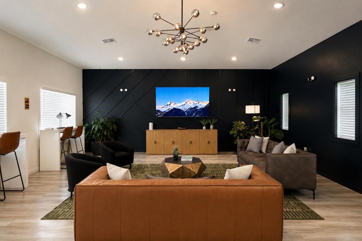 A modern living room featuring two stylish sofas, one brown leather and the other dark fabric, a geometric coffee table, and a flat-screen TV mounted on a black accent wall. There are plants, a bar area with stools, and large windows allowing natural light to fill the space. The decor is contemporary and inviting.