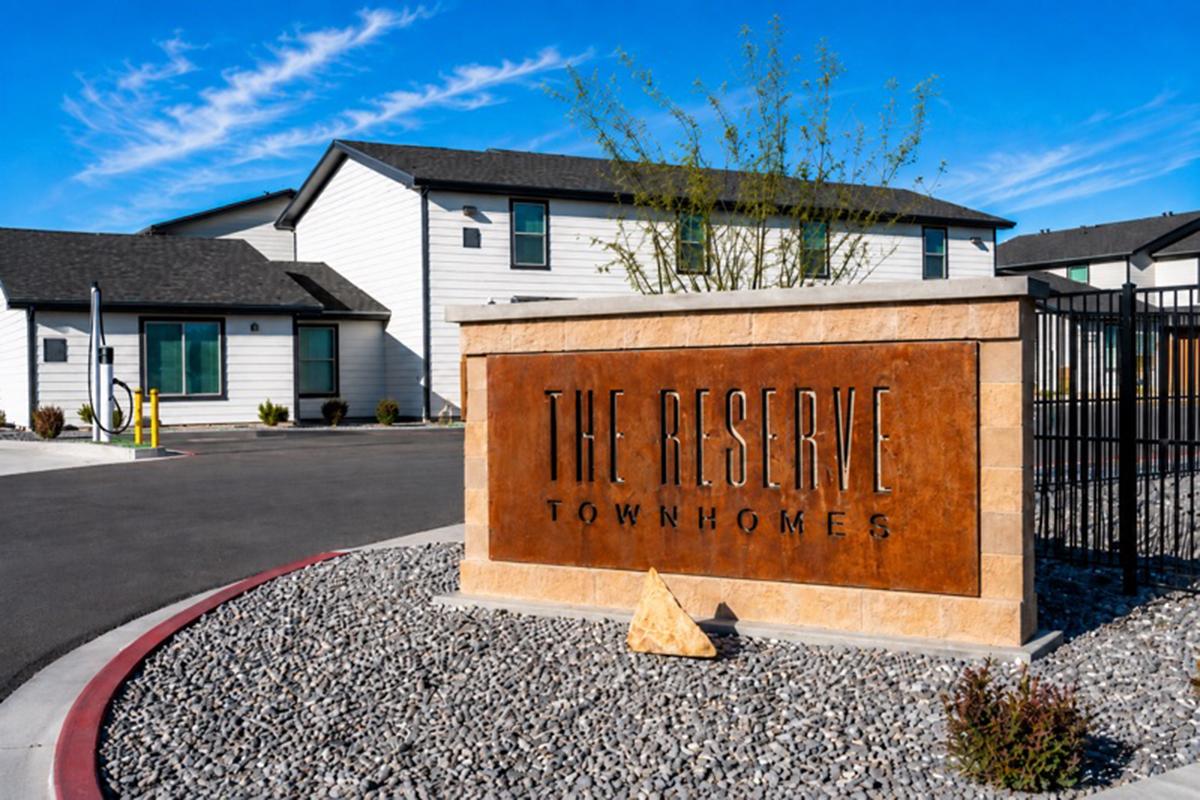 A modern entry sign for "The Reserve Townhomes" displayed prominently at the entrance of a residential community. The sign features a textured metallic finish with landscaping and apartment buildings visible in the background under a clear blue sky.
