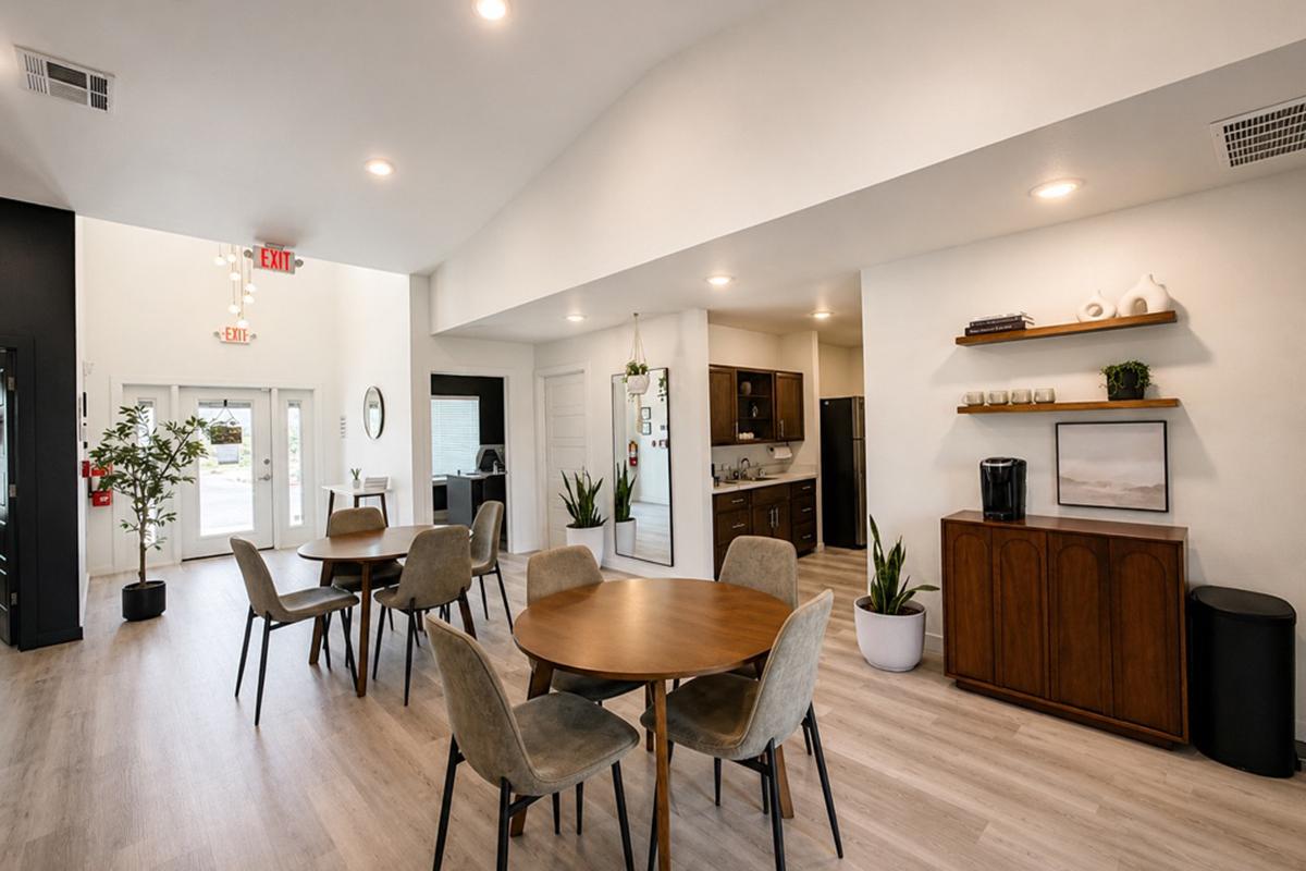 A modern communal space featuring a round wooden dining table surrounded by chairs, a kitchenette with dark cabinets, and a decorative sideboard. Potted plants add greenery, and large windows allow natural light to fill the area, creating a welcoming atmosphere.