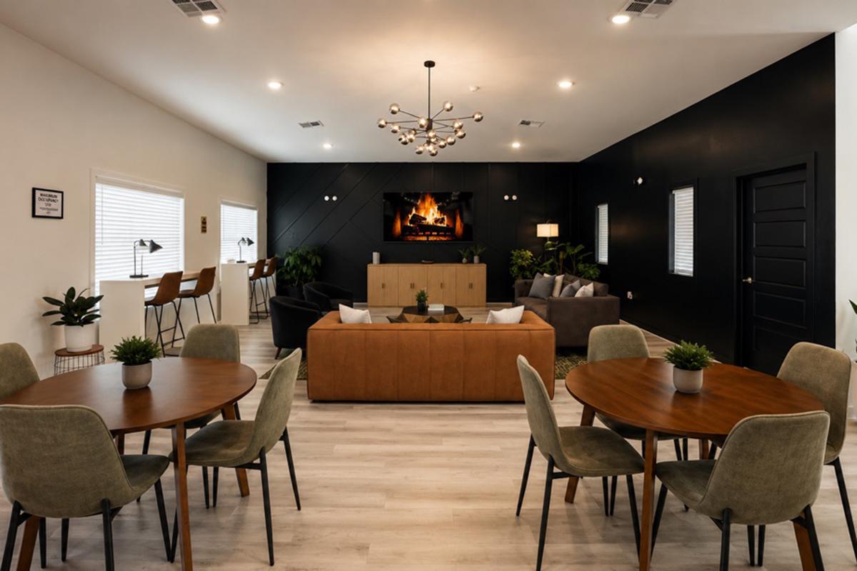 Modern living space featuring a cozy seating area with a fireplace, dark accent wall, and decorative plants. There are two round dining tables surrounded by upholstered chairs, and stylish lighting fixtures. The space has an inviting, contemporary design with warm colors and a blend of textures.