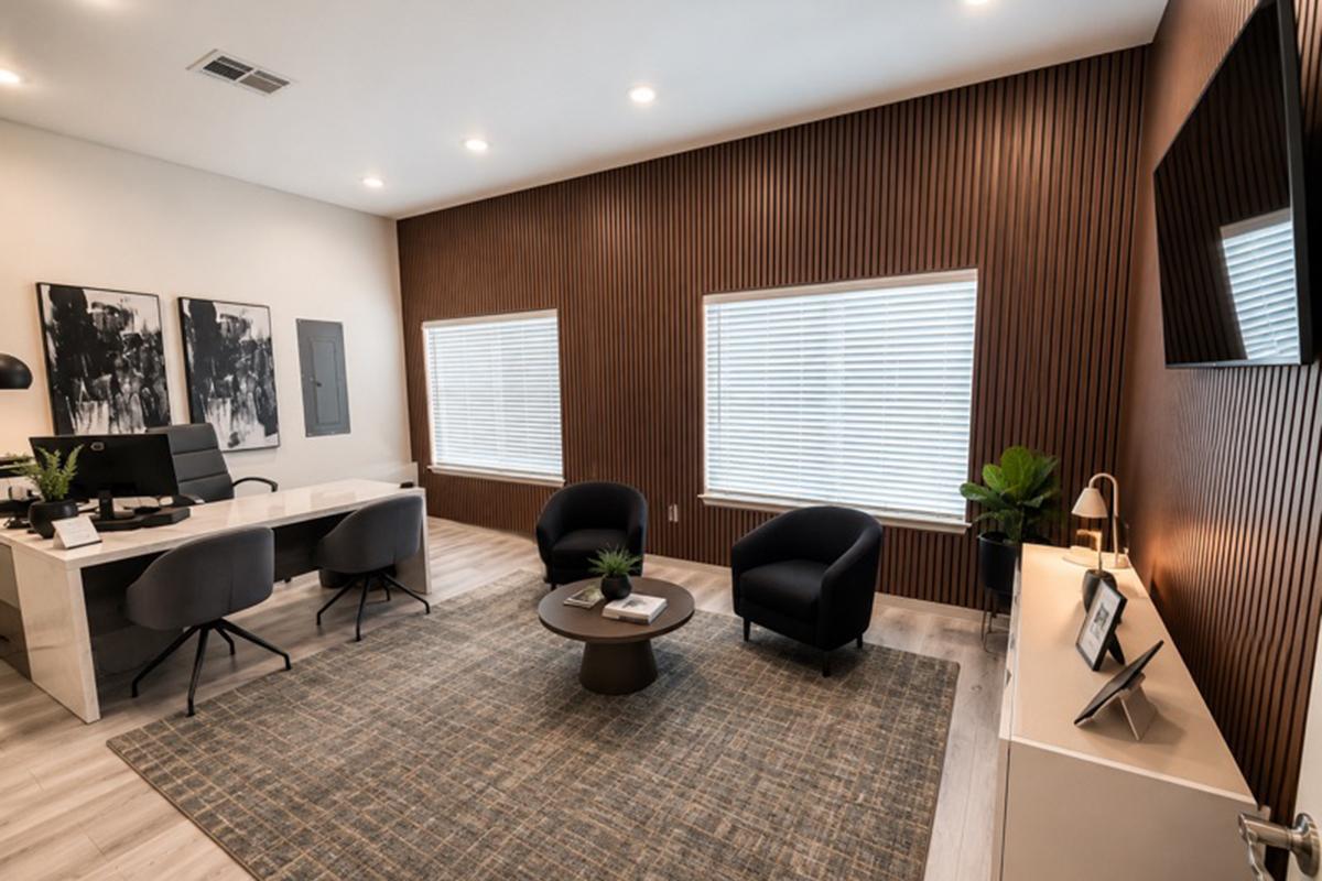A modern office interior featuring a white desk and ergonomic chairs. Two black armchairs sit near a round coffee table on a patterned rug. The walls have decorative wood paneling, and large windows with blinds allow natural light to fill the space. Decorative plants enhance the contemporary aesthetic.