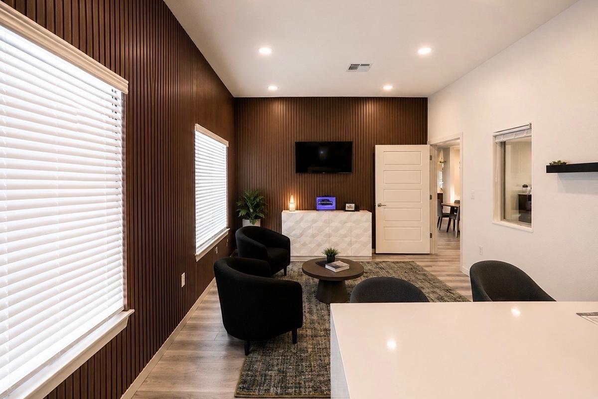 Modern office interior featuring a warm wooden accent wall, sleek furniture including black chairs, a light-colored accent table, and a TV mounted on the wall. Natural light enters through two windows with blinds. The space is stylishly decorated with minimalistic touches, creating a welcoming atmosphere.