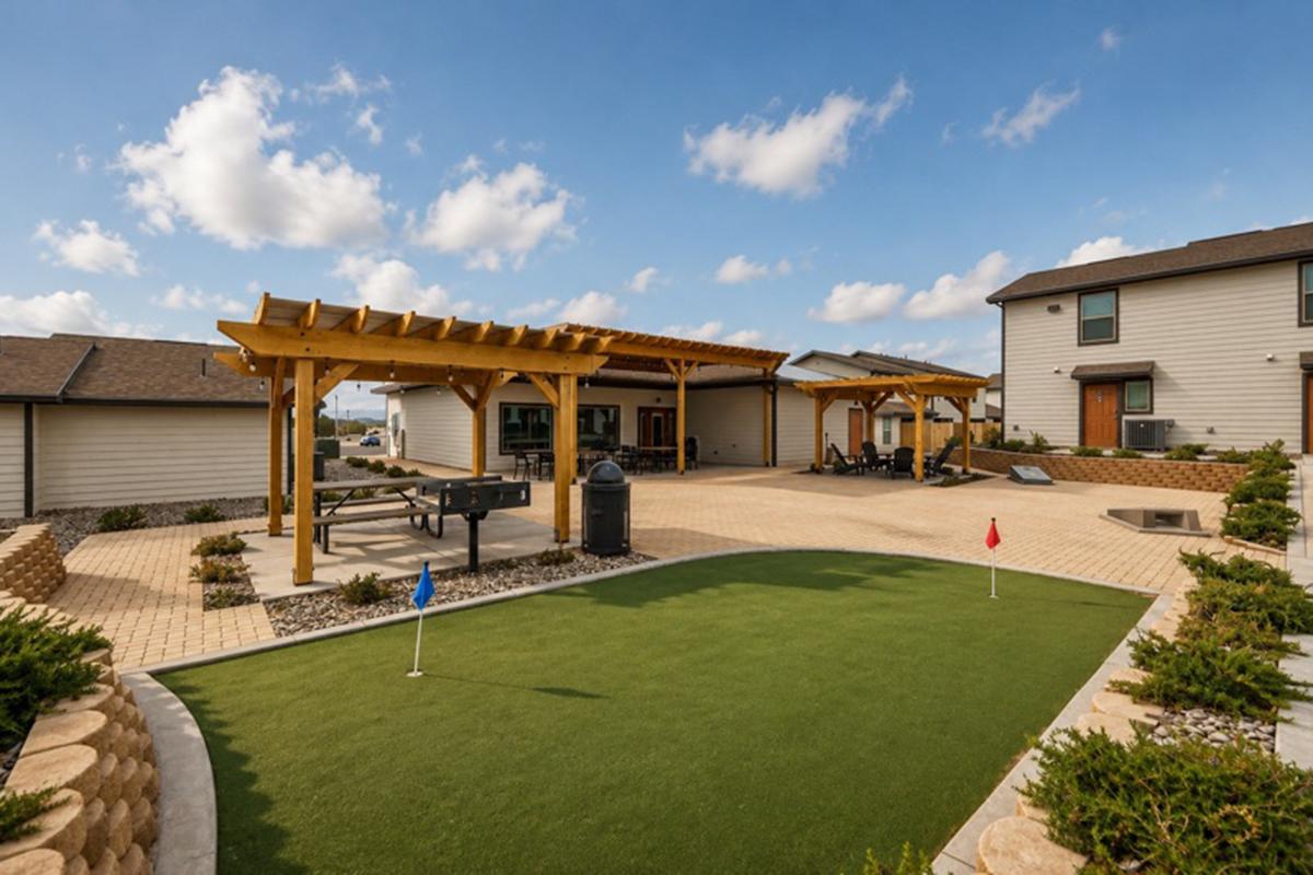 A landscaped outdoor space featuring a putting green with two flags, shaded pergolas, seating areas, and modern houses in the background under a blue sky with scattered clouds. The area is designed for leisure and socializing.