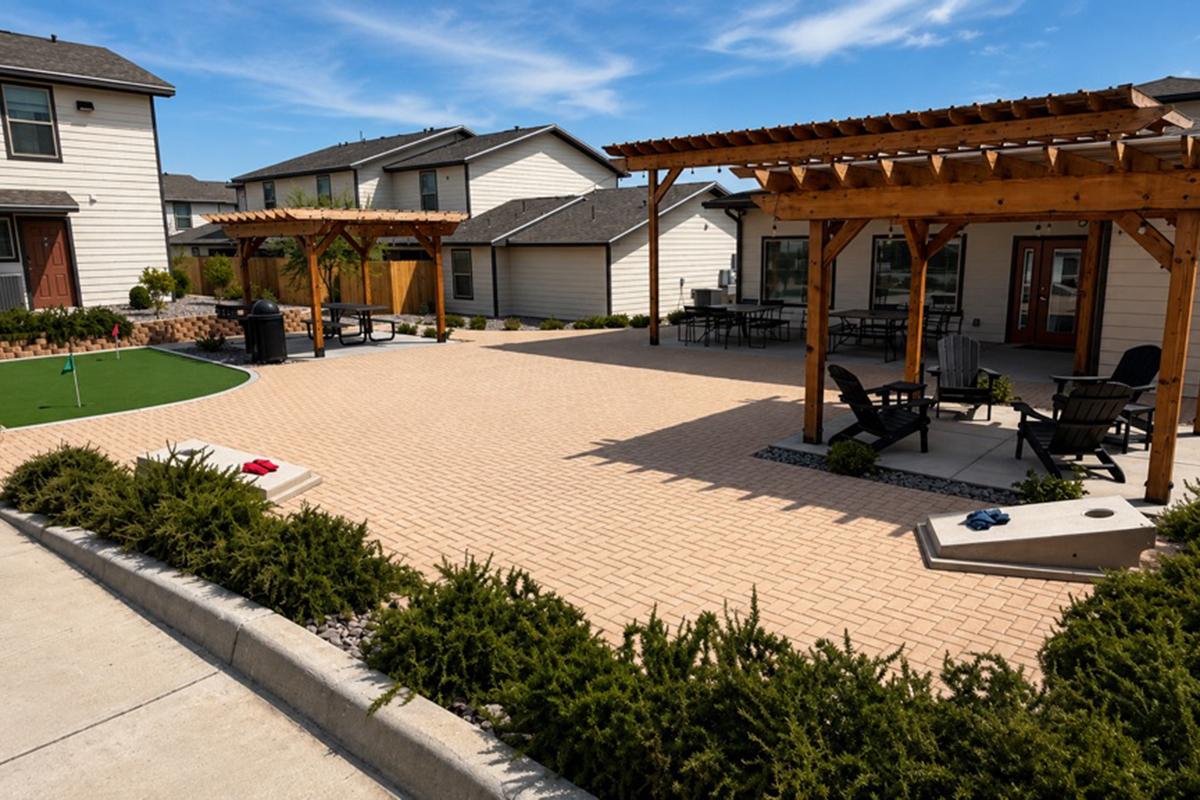Outdoor area featuring a spacious patio with patterned paving, surrounded by homes. There are wooden pergolas providing shade, outdoor seating with black chairs, a putting green, and a small garden area with low shrubs. The sky is clear with a few clouds.