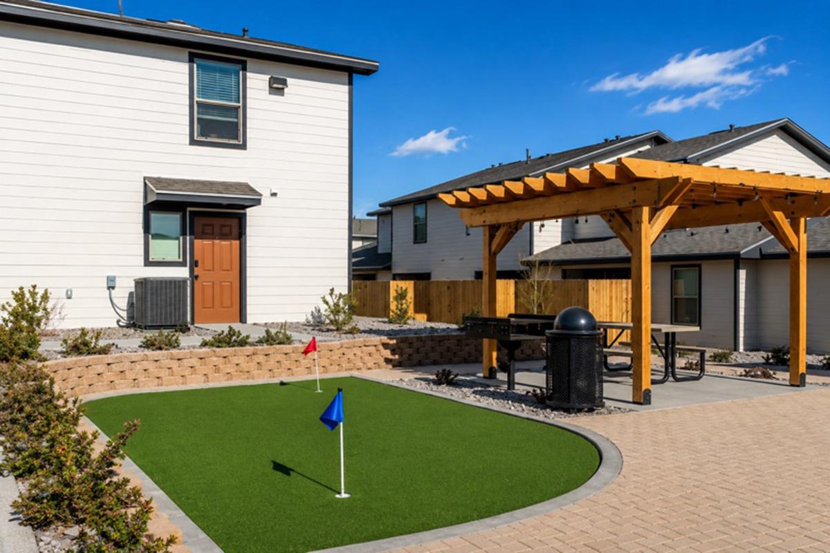 A residential backyard featuring a small putting green with a red and blue flag, a patio area with a picnic table, a grill, and a pergola. The house has a brown door and a well-maintained garden with low shrubs. Clear blue skies and modern homes are visible in the background.