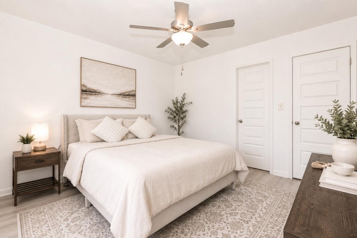 A bright and airy bedroom featuring a queen-sized bed with light bedding, two decorative pillows, and a textured headboard. A wooden nightstand holds a lamp, while a potted plant adds greenery. The room has a ceiling fan and white walls, with artwork above the bed and a neutral rug on the floor. Two closed doors are visible.