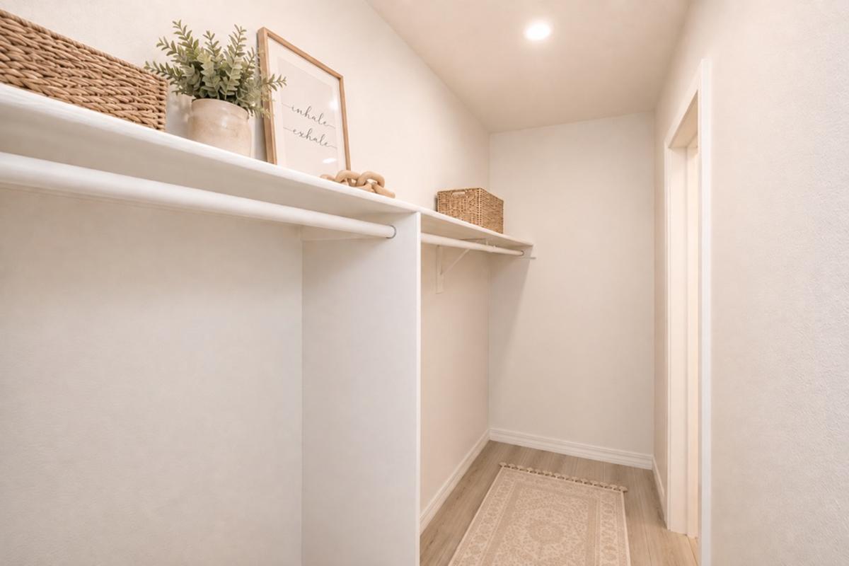 A clean, minimalist hallway featuring white walls and a light wooden floor. There are shelves on one side holding decorative baskets and a small plant. A framed artwork is mounted on the wall, and a textured rug lies on the floor, adding warmth to the space. A door is visible at the end of the hallway.