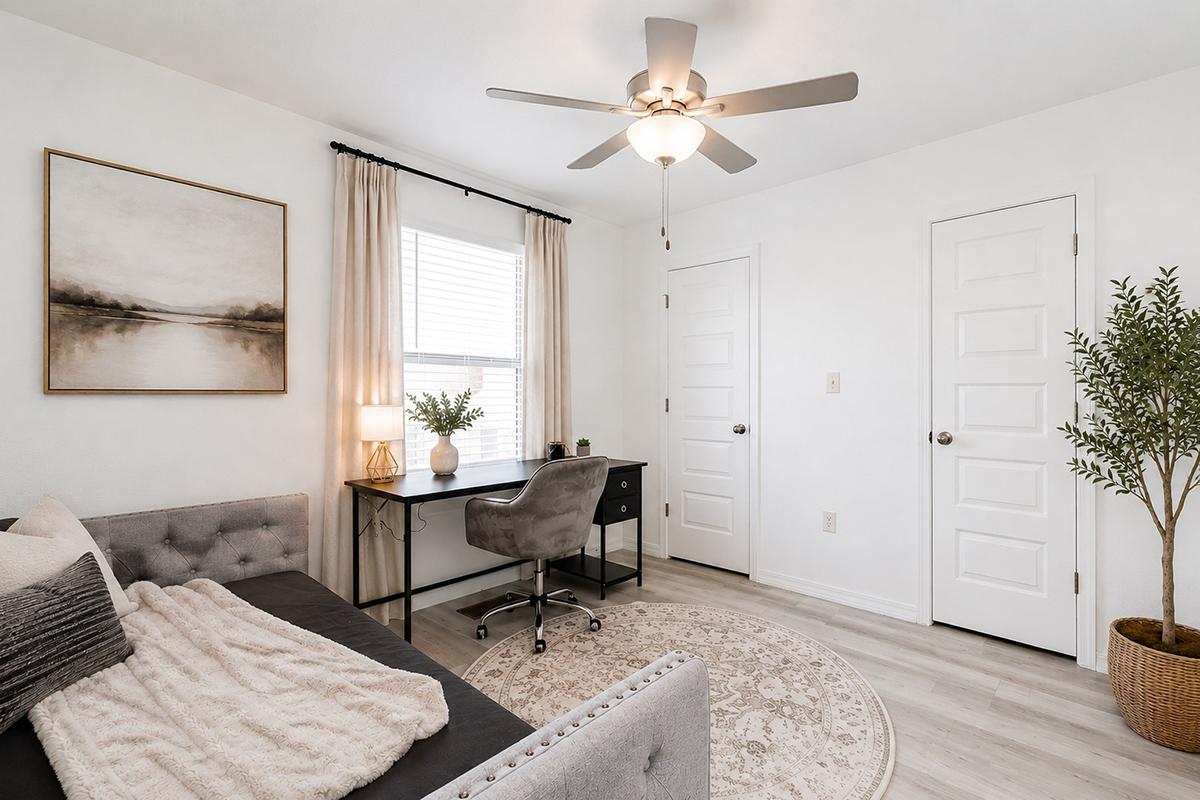 A cozy, modern room featuring a gray sofa with a soft throw, a small desk with a stylish chair, and a decorative plant. Natural light streams in through a window dressed with sheer curtains. The walls are painted white, and there is a round area rug on the floor, creating a warm and inviting atmosphere.