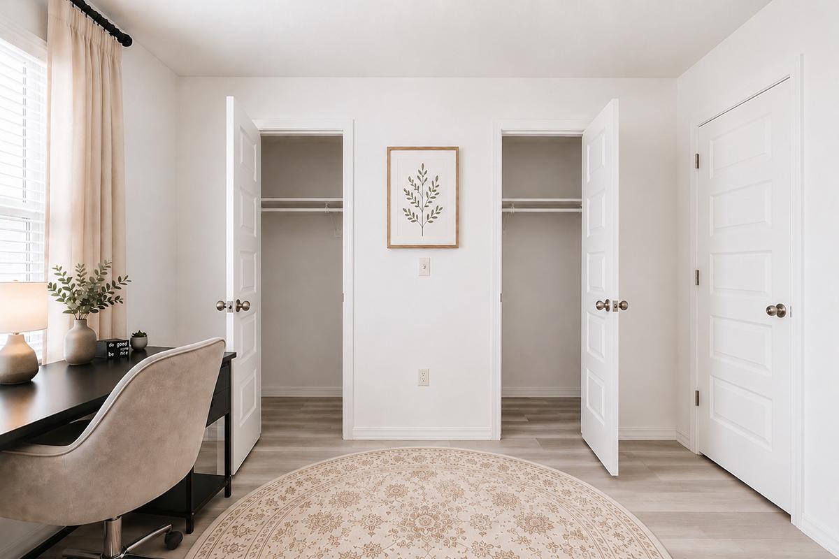 A bright, minimalistic room featuring two open closet doors on either side, a desk with a chair and a lamp on the left, and a round area rug on the floor. A framed plant illustration is on the wall, and light-colored curtains adorn the window, creating a serene and organized atmosphere.