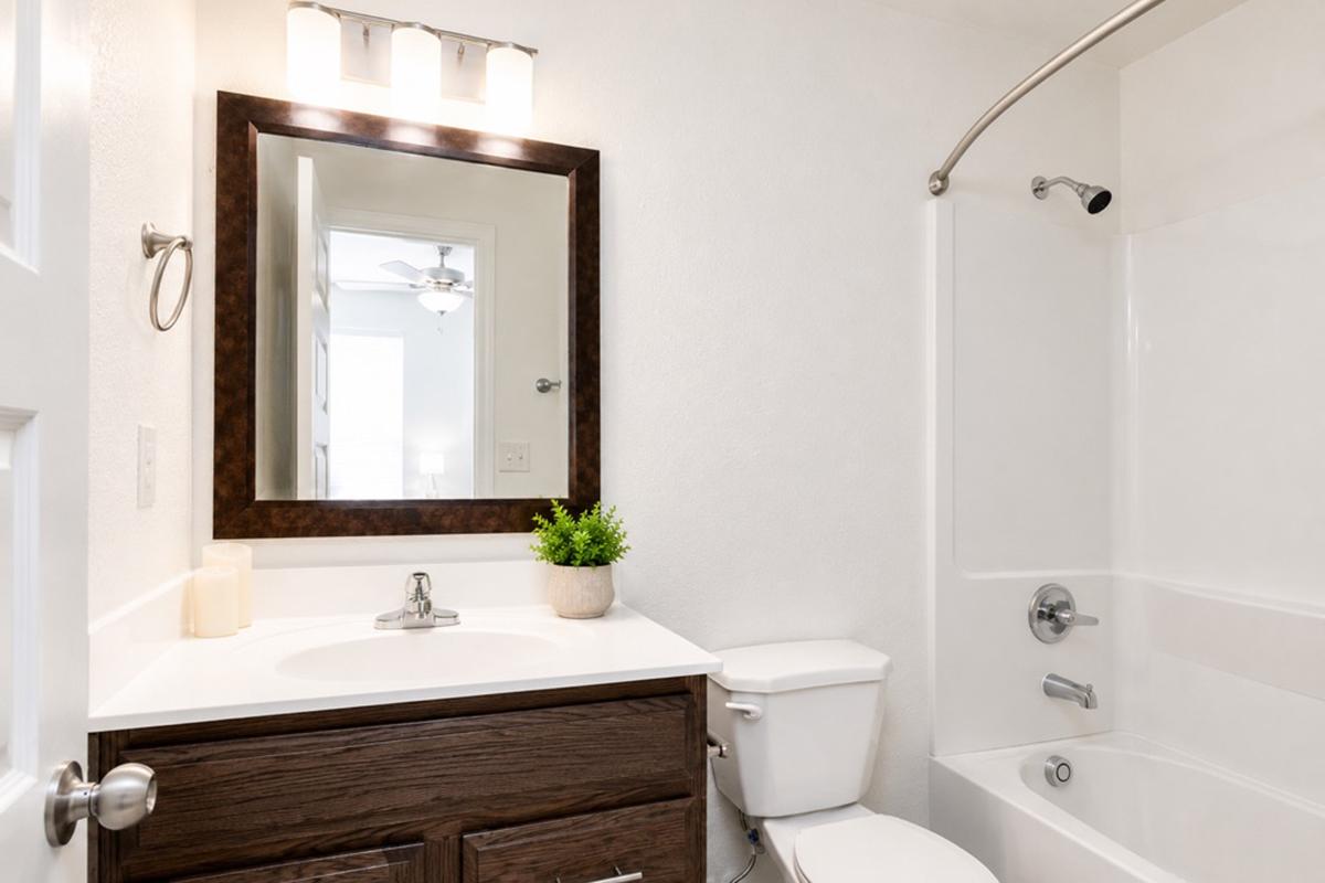 A well-lit bathroom featuring a white and brown color scheme. The vanity has a modern sink and faucet, with a small potted plant on top. A large mirror is above the sink, and there is a bathtub with a shower curtain on the right. The wall is painted white, and a light fixture is mounted above the mirror.