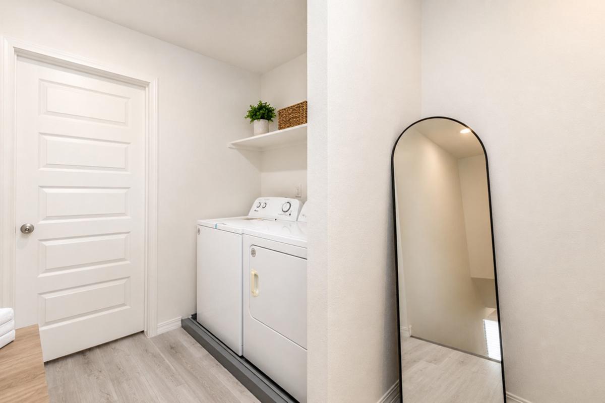 A bright and clean laundry room featuring a white washing machine and dryer side by side. There is a shelf above with a small plant and a basket. A full-length mirror is positioned against the wall, reflecting the light and creating an open feel, with neutral-colored walls and light wood flooring.
