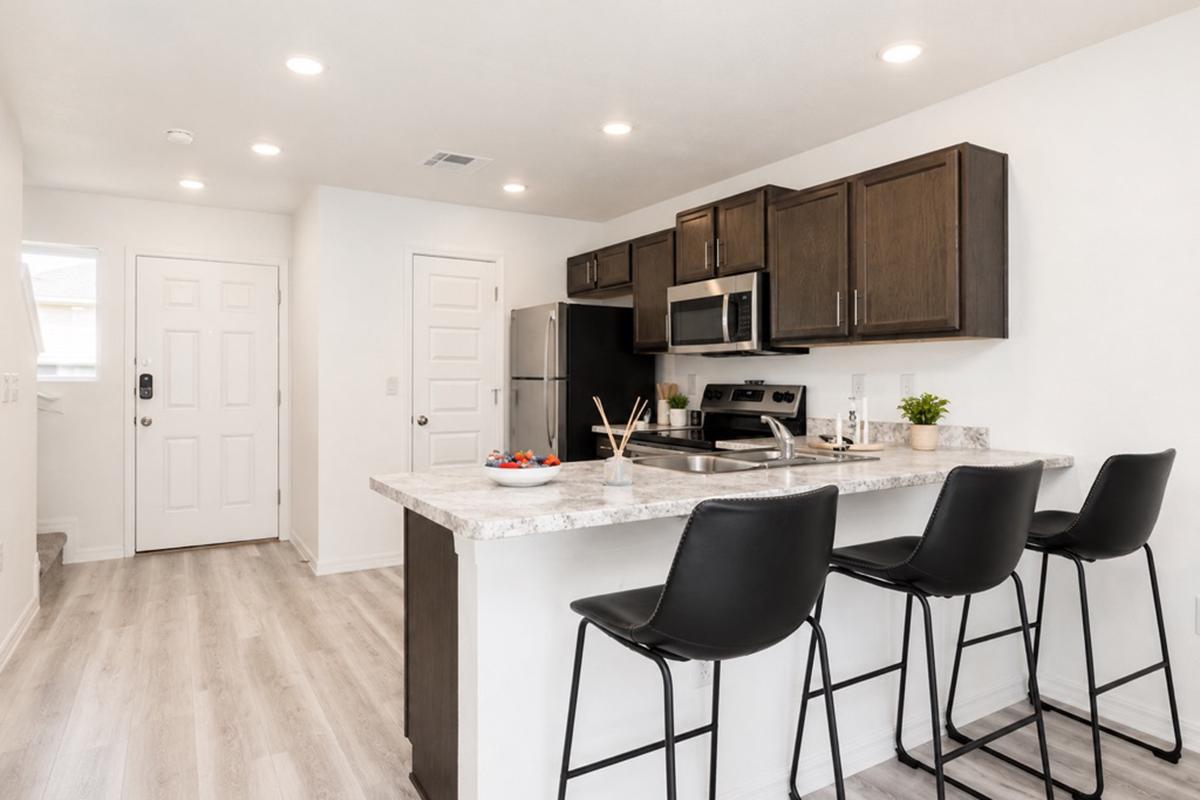 Modern kitchen featuring a granite countertop with three black bar stools, stainless steel appliances including a refrigerator and stove, light wood flooring, and bright lighting fixtures. An entry door is visible on the left side, enhancing the open and inviting atmosphere of the space.
