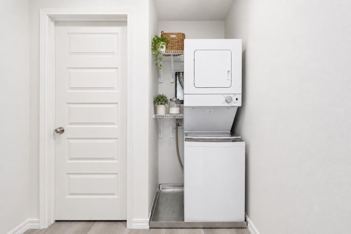 A compact laundry area featuring a stacked washer and dryer set against a white wall. There is a closed white door on the left and shelves above the appliances with potted plants and a woven basket, creating a neat and organized space.
