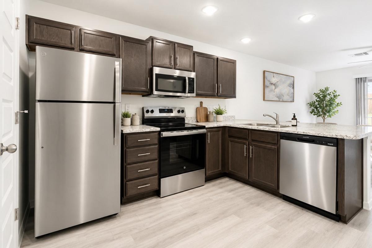 Modern kitchen featuring stainless steel appliances, including a refrigerator, microwave, oven, and dishwasher. The cabinetry is dark wood with a light countertop. Decorative plants are on the counter, and there's a light-filled atmosphere with a window and neutral-colored walls.
