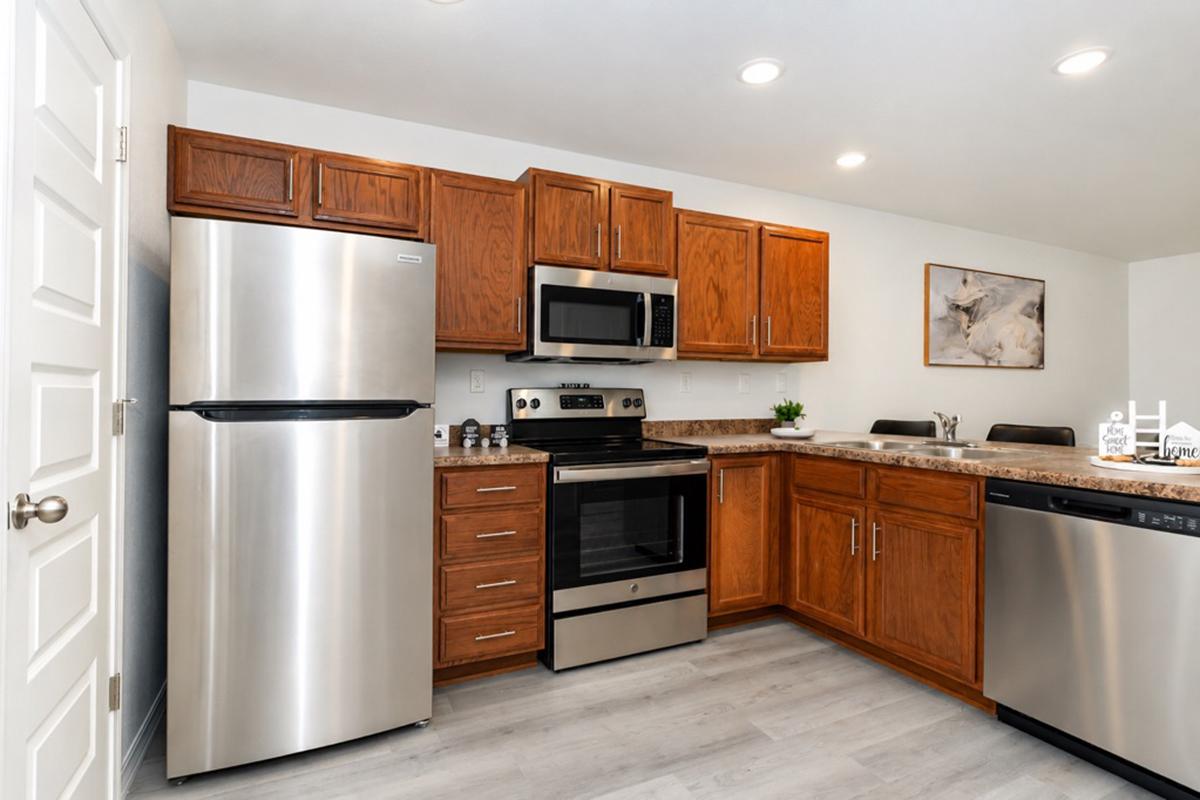 Modern kitchen featuring stainless steel appliances, including a refrigerator, oven, and dishwasher. The cabinetry is wood-toned, with a granite countertop. The kitchen is well-lit with recessed lighting and includes a dining area with two chairs. A piece of art is displayed on the wall.