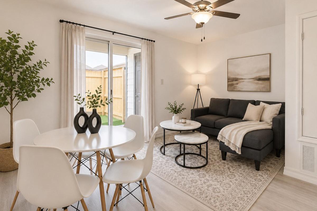 A modern living room featuring a grey sectional sofa with cushions, a round side table, and a coffee table. A white dining table with four chairs is in the foreground. Large windows allow natural light, and a potted plant adds a touch of greenery. Neutral tones and simple decor create a cozy atmosphere.