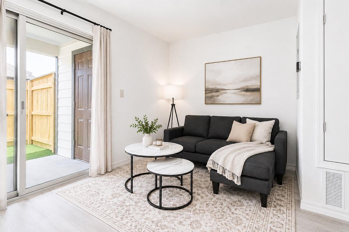 A modern living room featuring a dark gray sofa with decorative pillows, a round coffee table with a marble top, and a lamp. There’s a large sliding door leading to a small outdoor area, with a beige area rug on light wooden flooring and a wall art piece depicting a serene landscape. Greenery is displayed on the table.