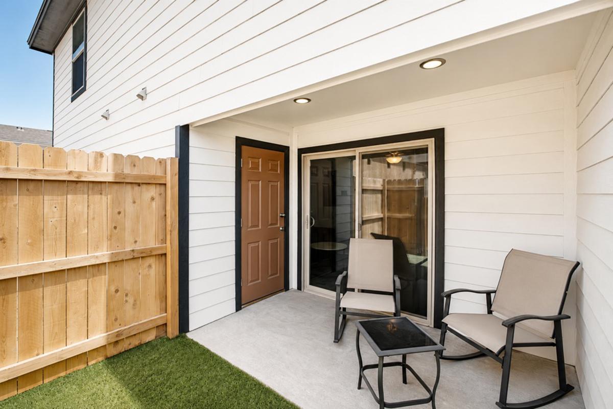 A small outdoor patio featuring two beige chairs and a black table, framed by a wooden fence. The patio is attached to a modern-looking house with a sliding glass door, allowing access to the indoor area. The space is well-lit, giving a cozy and inviting atmosphere.