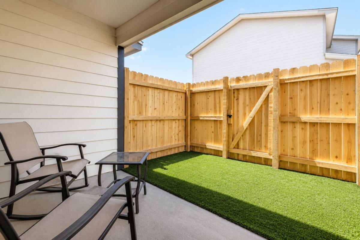 A small outdoor patio area with two chairs and a glass table on synthetic grass. A wooden fence encloses the space, providing privacy. Bright blue skies are visible above, and a house can be seen in the background. The overall setting offers a cozy and inviting atmosphere.