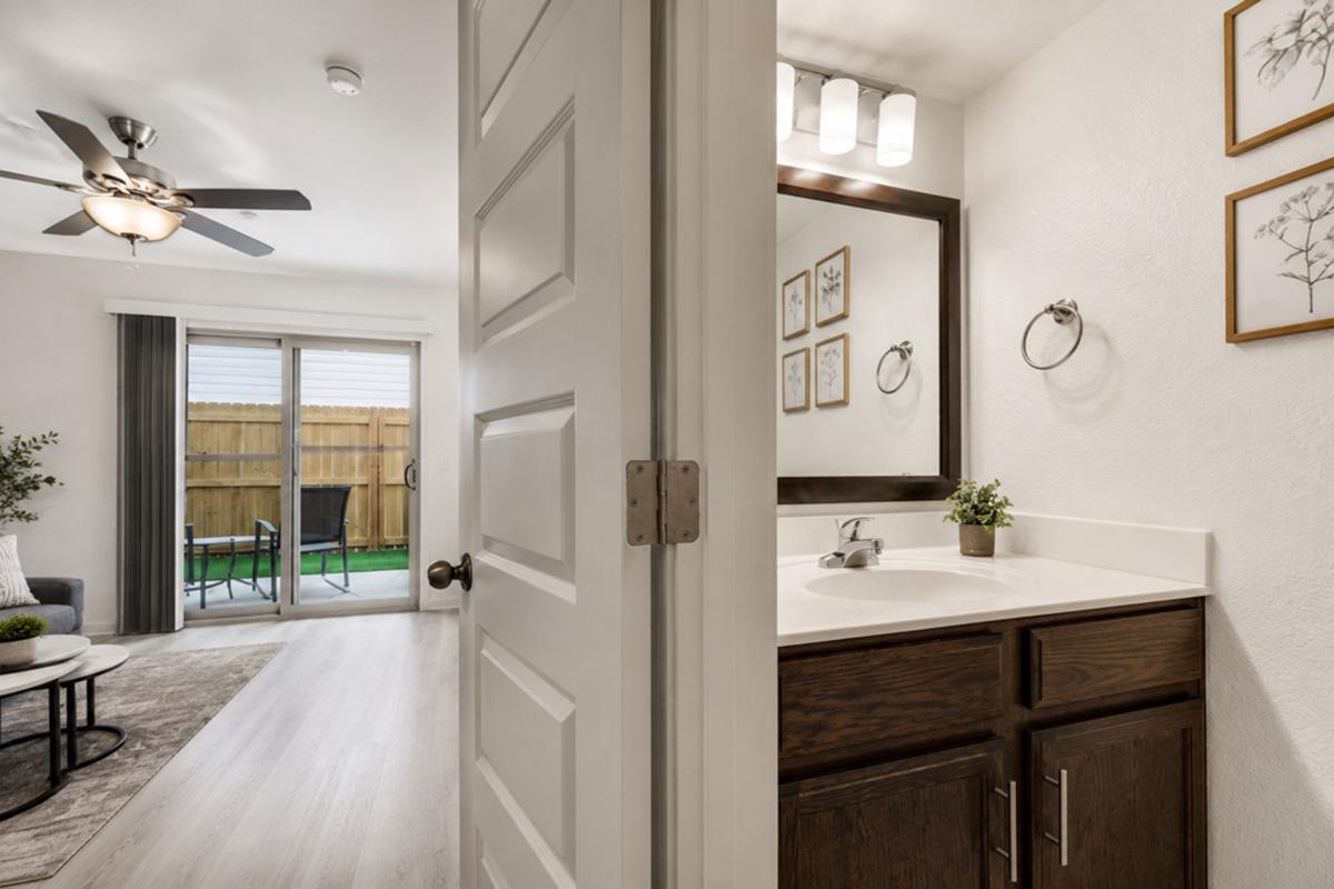 View of a bathroom with a sink and mirror, featuring dark wood cabinetry. A door opens to a living area with a patio outside, visible through sliding glass doors. The space includes a ceiling fan and decorative wall art, creating a cozy and modern atmosphere.