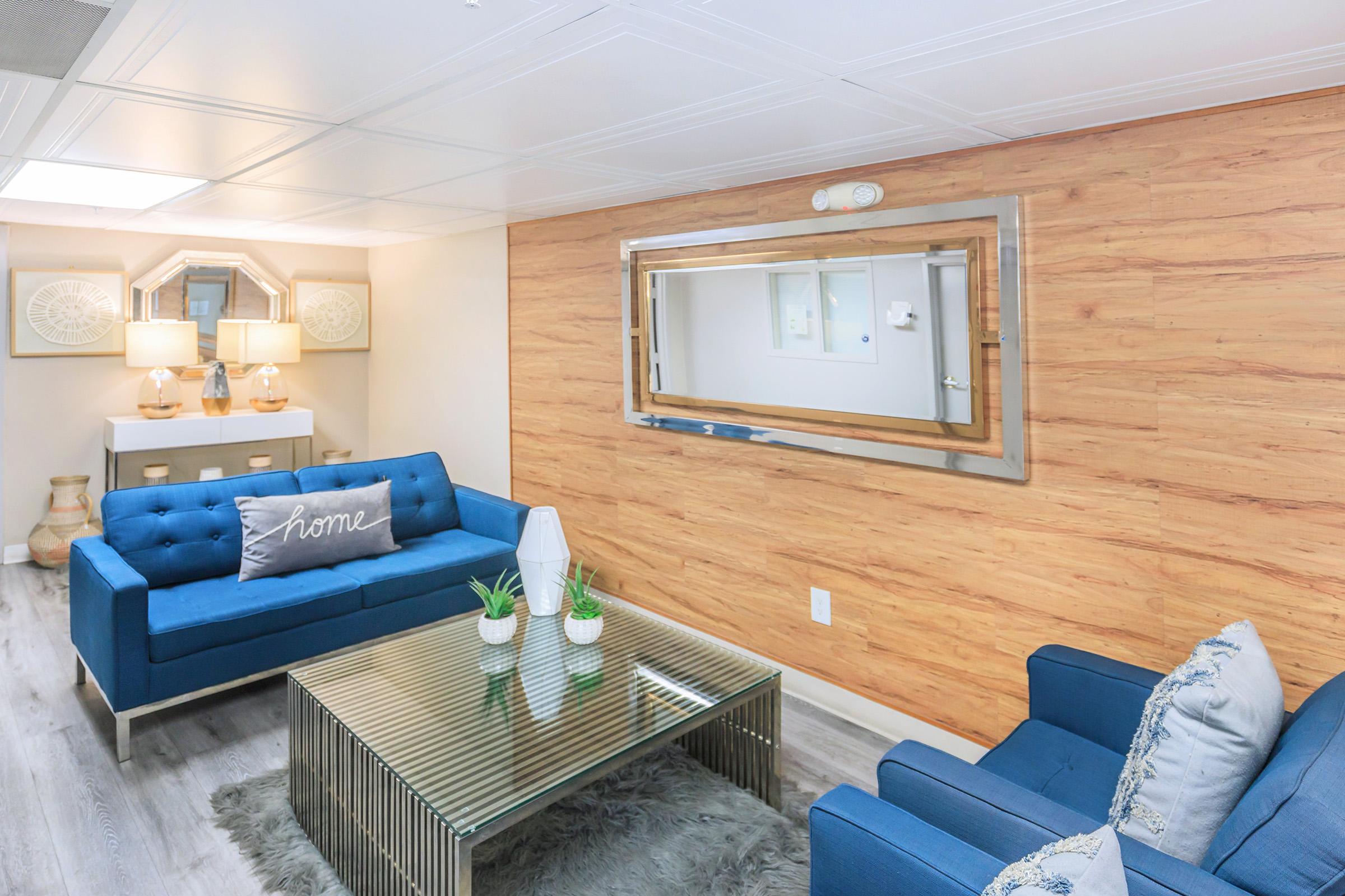A cozy living area featuring two blue sofas, a glass coffee table, and decorative plants. The wall is adorned with wood paneling and a large mirror, while two lamps provide warm lighting. A soft "home" pillow adds a touch of comfort to the space.