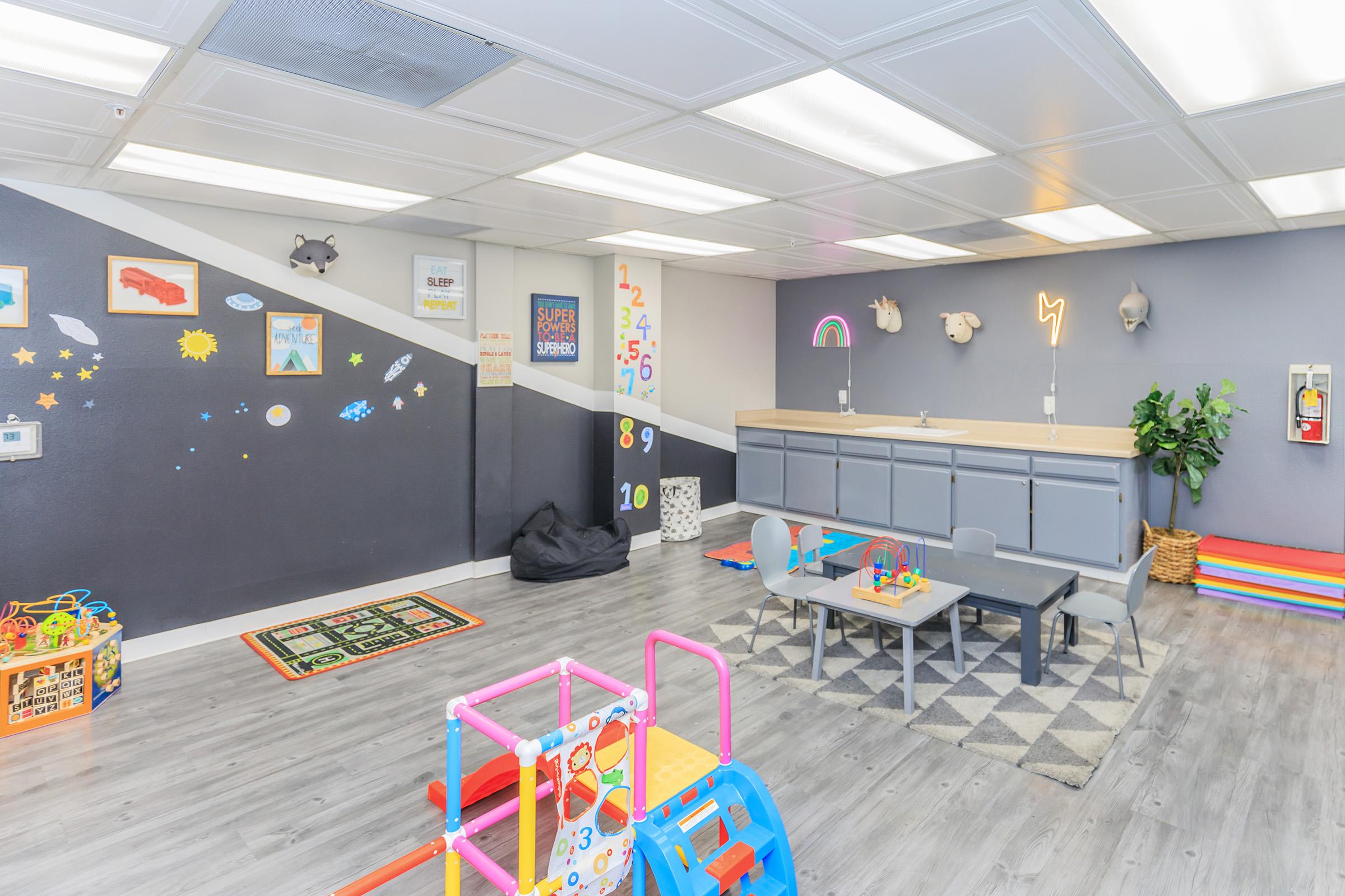A bright and colorful children's playroom featuring a variety of toys, a small table with chairs, a play kitchen area, and decorative wall art, including numbers and shapes. Soft flooring and a cozy atmosphere create an inviting space for kids to play and learn.