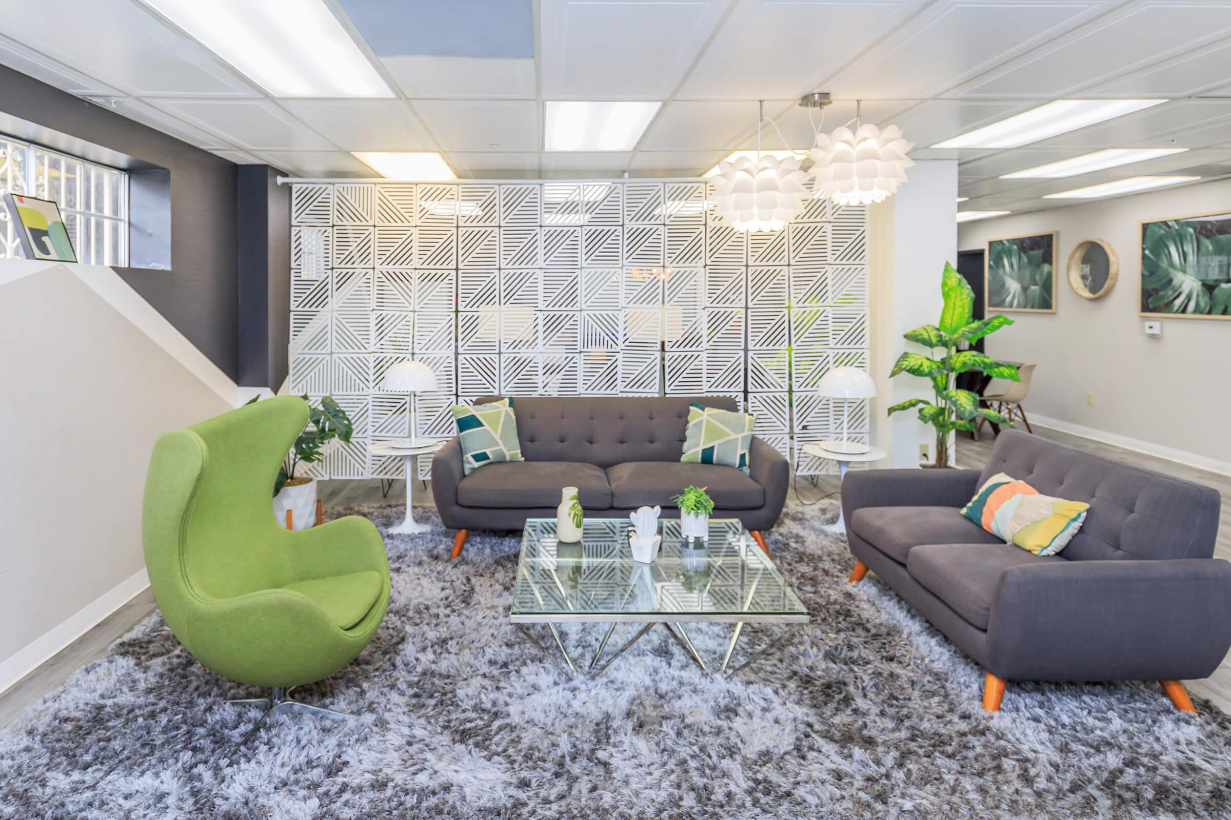 A modern and stylish lounge area featuring a gray sofa and a green, egg-shaped chair. There is a glass coffee table on a plush gray rug, complemented by decorative pillows. The backdrop includes a white geometric-patterned wall and leafy plants, creating a welcoming and contemporary atmosphere.