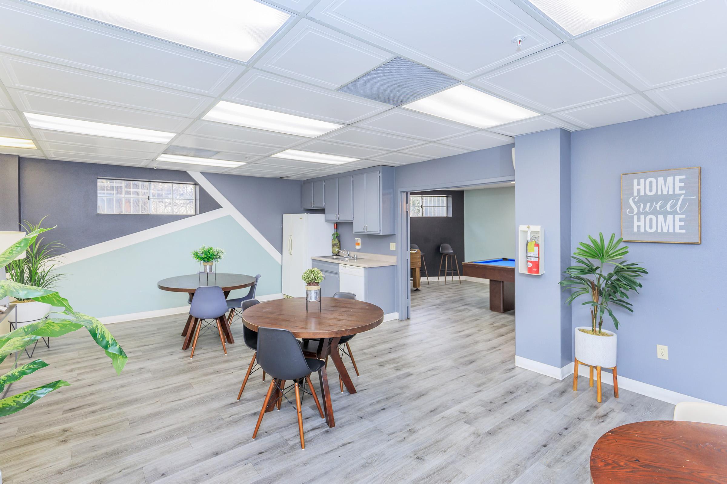 A modern shared space featuring several wooden round tables with chairs, a kitchenette area with a fridge, and light blue walls. There are potted plants and a wall sign that reads "Home Sweet Home." Natural light enters through the windows, creating a welcoming atmosphere.