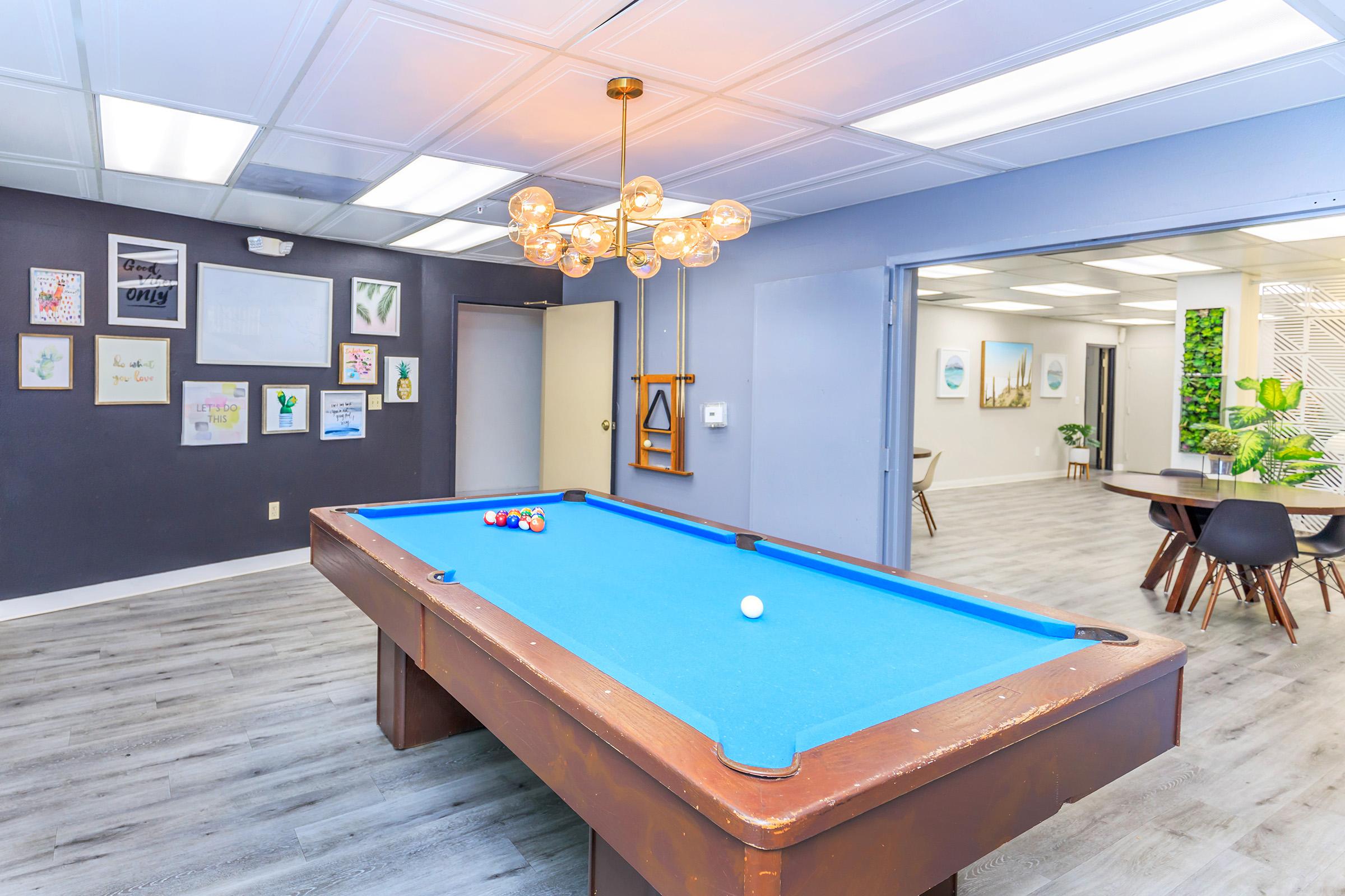 A spacious room featuring a blue felt pool table with a cue ball and colorful balls nearby. The walls are adorned with framed artwork and a modern chandelier hangs from the ceiling. In the background, another room can be seen with a dining table and greenery. The flooring is light wood, enhancing the bright atmosphere.