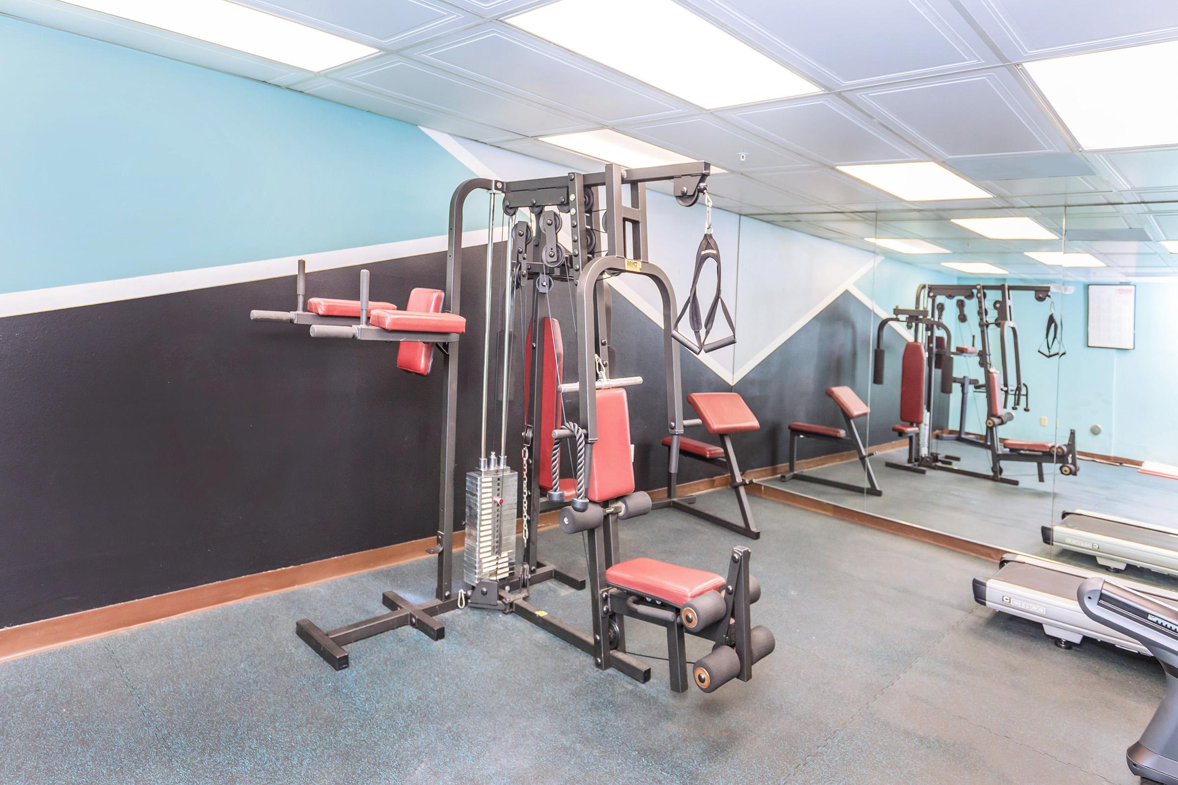 A gym featuring a multi-station exercise machine with red seating, various attachments, and weights. In the background, mirrors reflect the equipment and blue and black wall colors, creating a spacious workout environment.