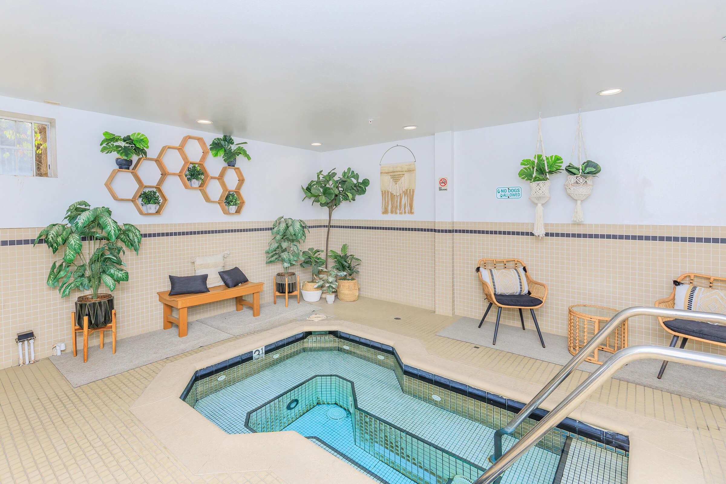 Indoor spa area featuring a small hot tub surrounded by potted plants and decorative hexagonal wall shelves. Furnished with a couple of chairs and a small table, the space has a calming ambiance with neutral colors and natural elements. Soft lighting enhances the relaxing atmosphere.