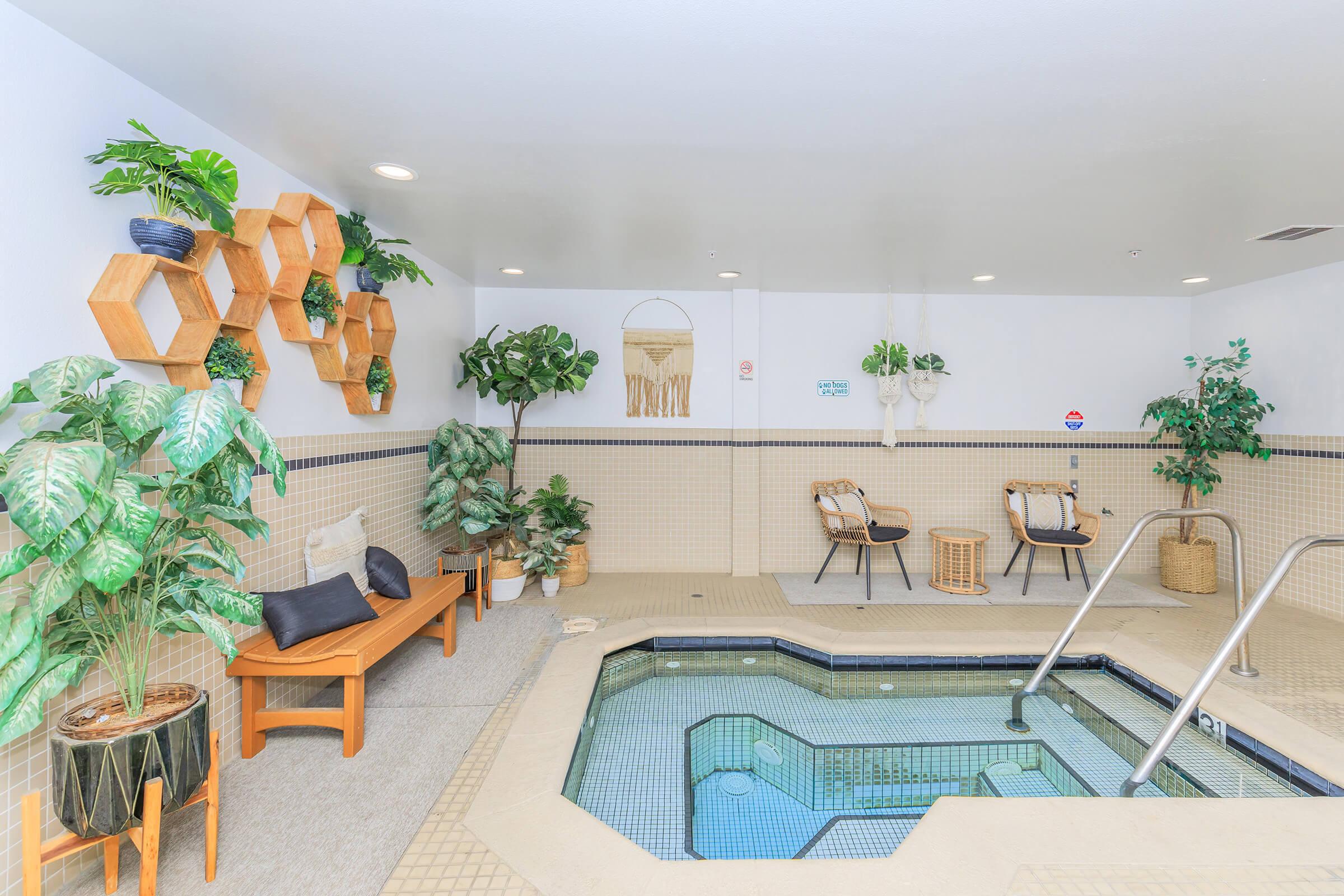 Indoor spa area with a hot tub surrounded by hexagonal wooden shelves and lush green plants. There are two woven chairs and a wooden bench for seating. The walls are decorated with light tiles, and natural light brightens the space, creating a calming atmosphere.