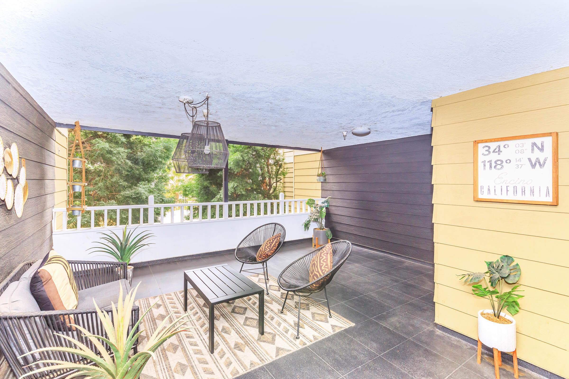 A modern patio with a cozy seating arrangement featuring two round chairs and a small table. The area is decorated with a patterned rug and potted plants. A framed sign displaying coordinates and the word "California" is mounted on the wall, while trees are visible outside through the railing.