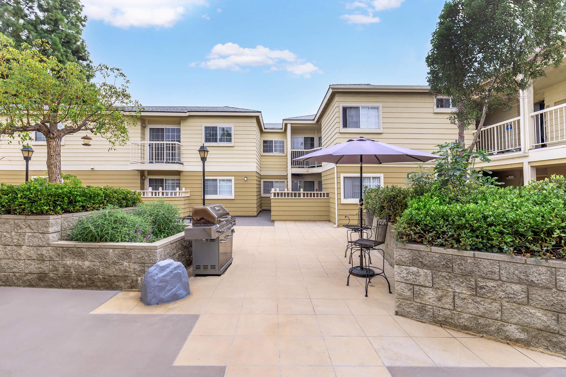 A landscaped courtyard featuring a stone wall, greenery, and a shaded patio area with a table and chairs. A barbecue grill is situated nearby, and there are two-story apartment buildings in the background under a clear sky.