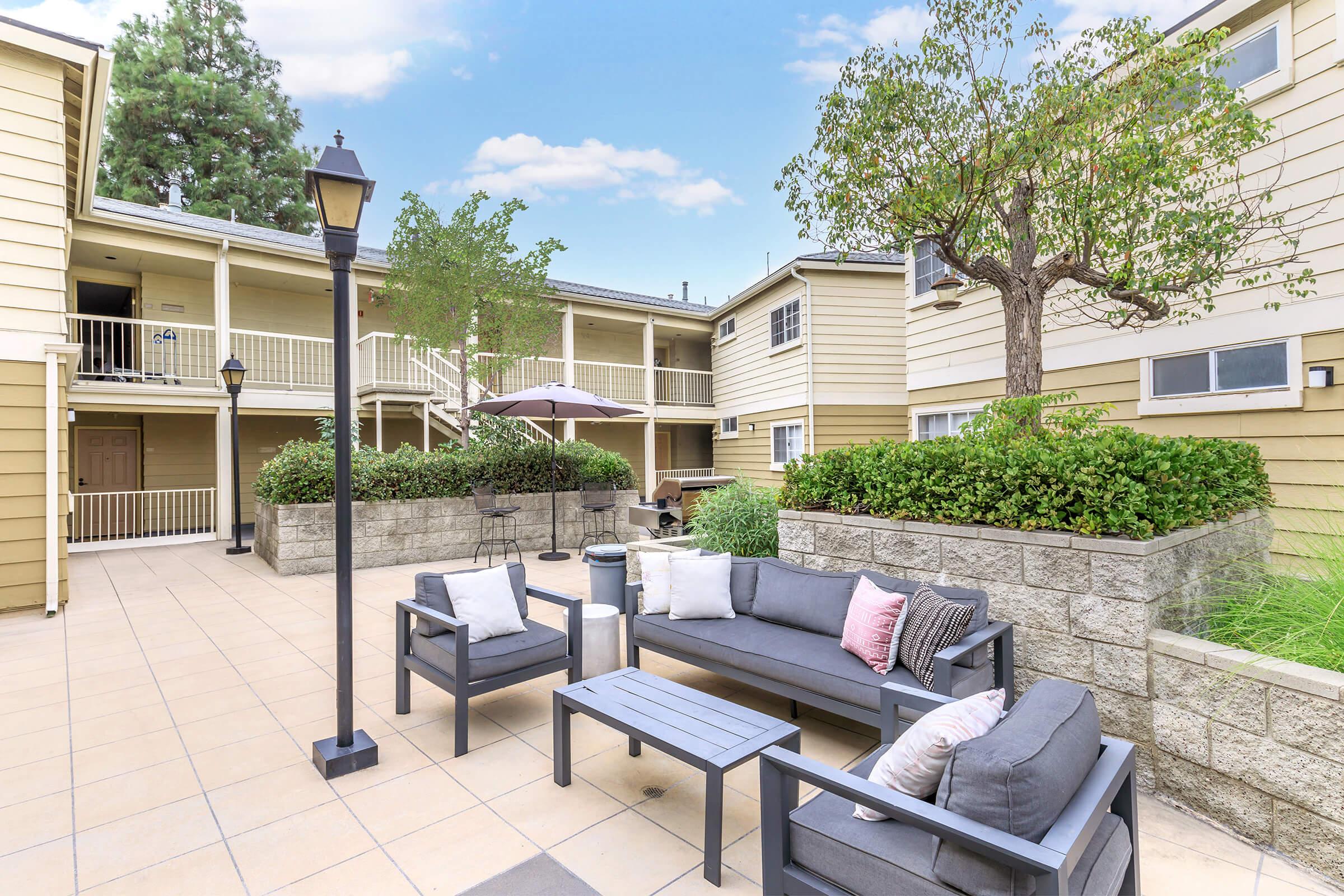 A courtyard area featuring modern outdoor furniture, including a gray sofa and coffee table, surrounded by potted greenery and light fixtures. In the background, there are two-story buildings with balconies and an umbrella over a seating area, all under a blue sky.