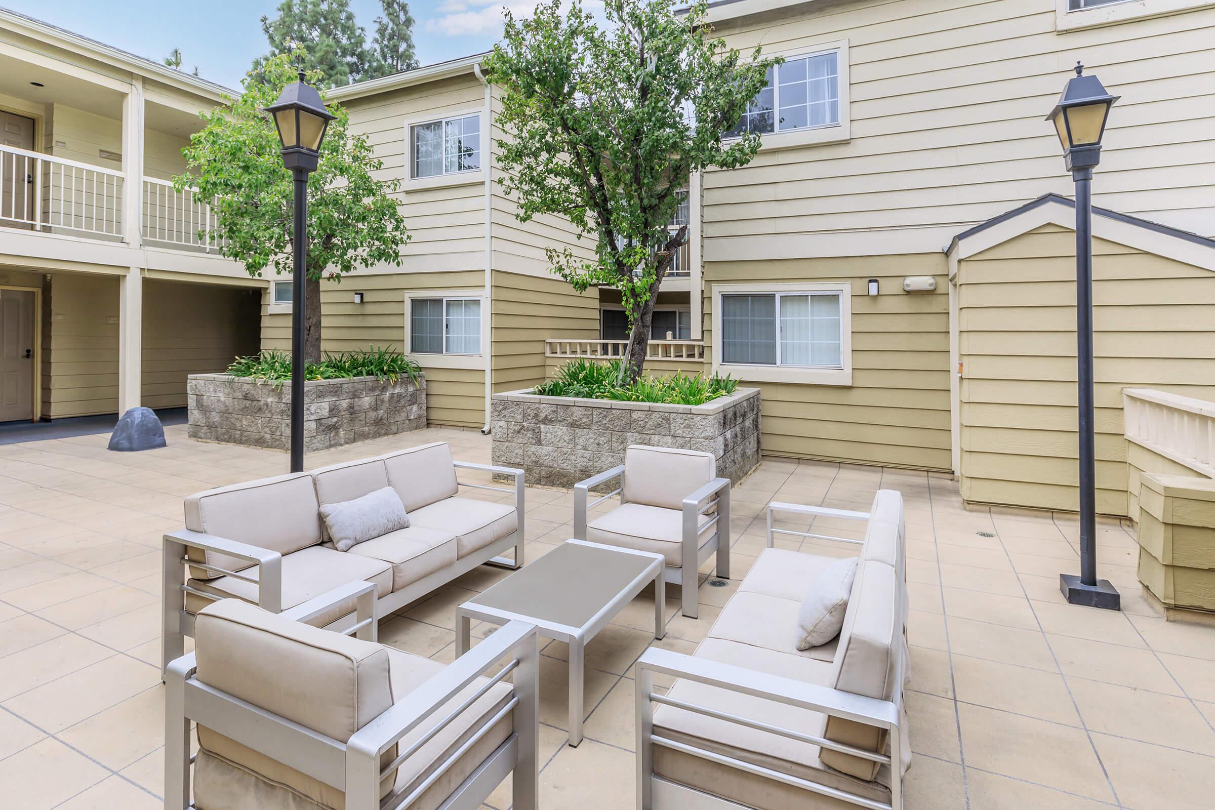 A cozy outdoor seating area featuring modern patio furniture including sofas, a coffee table, and a tree in the background. The surroundings are part of a residential building with light-colored siding and a small shed. There are also lampposts providing soft lighting.