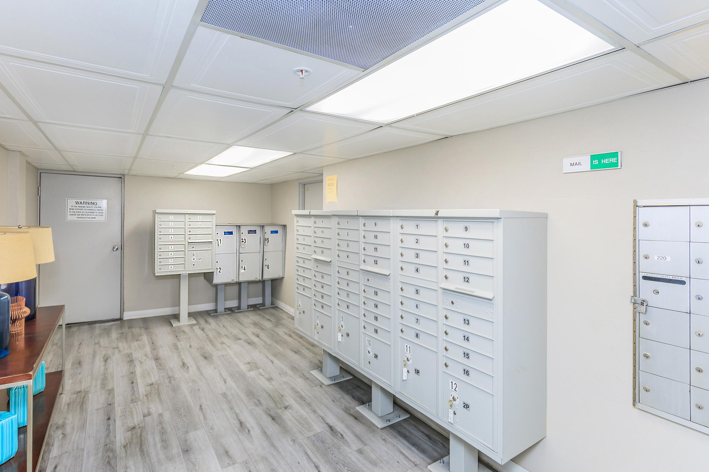 Interior view of a mailroom featuring several rows of gray mailboxes, a wooden table with a lamp on the left, and bright overhead lighting. There are signs on the walls, including a warning notice and a green label. The floor is covered with light-colored wood-like vinyl.
