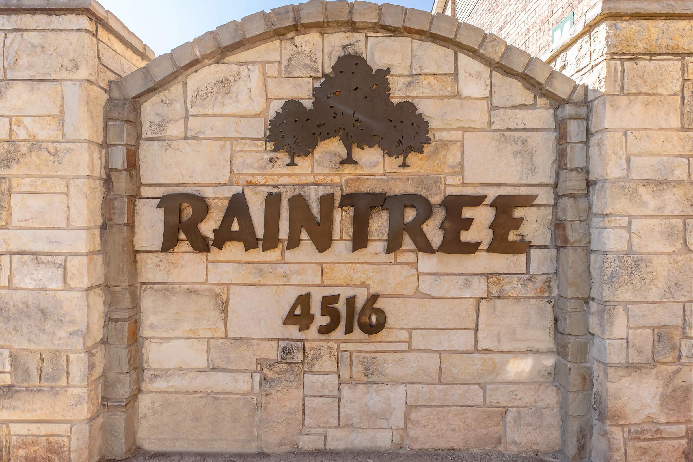 Stone archway sign displaying the word "RAINTREE" and the number "4516," with decorative tree silhouettes above the text. The arch and sign are made of textured stone, with a warm color tone.