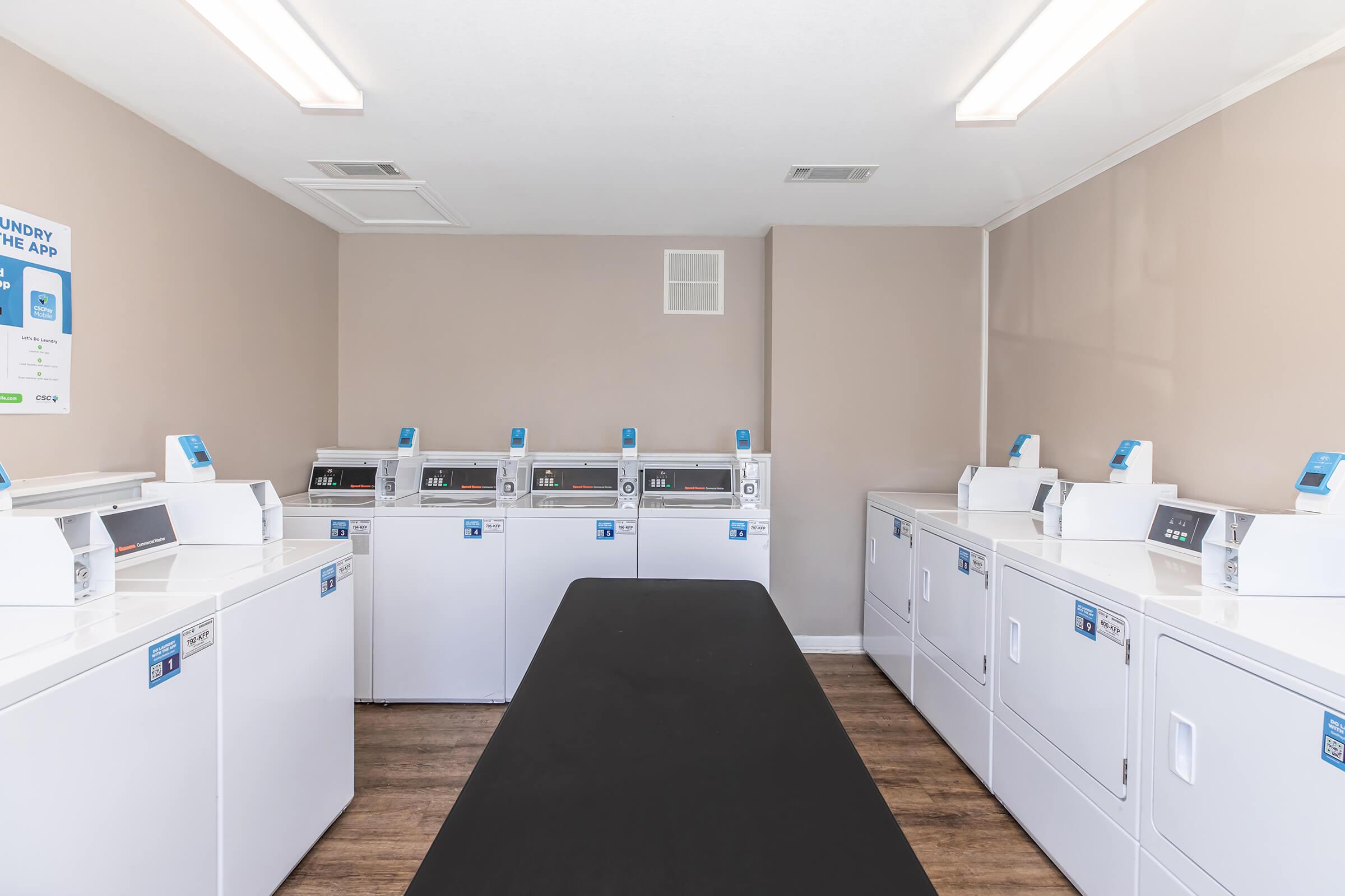 A clean and modern laundry room featuring several white washing machines and dryers aligned against a beige wall. In the center, there's a black folding table. Bright ceiling lights illuminate the space, creating a welcoming atmosphere.