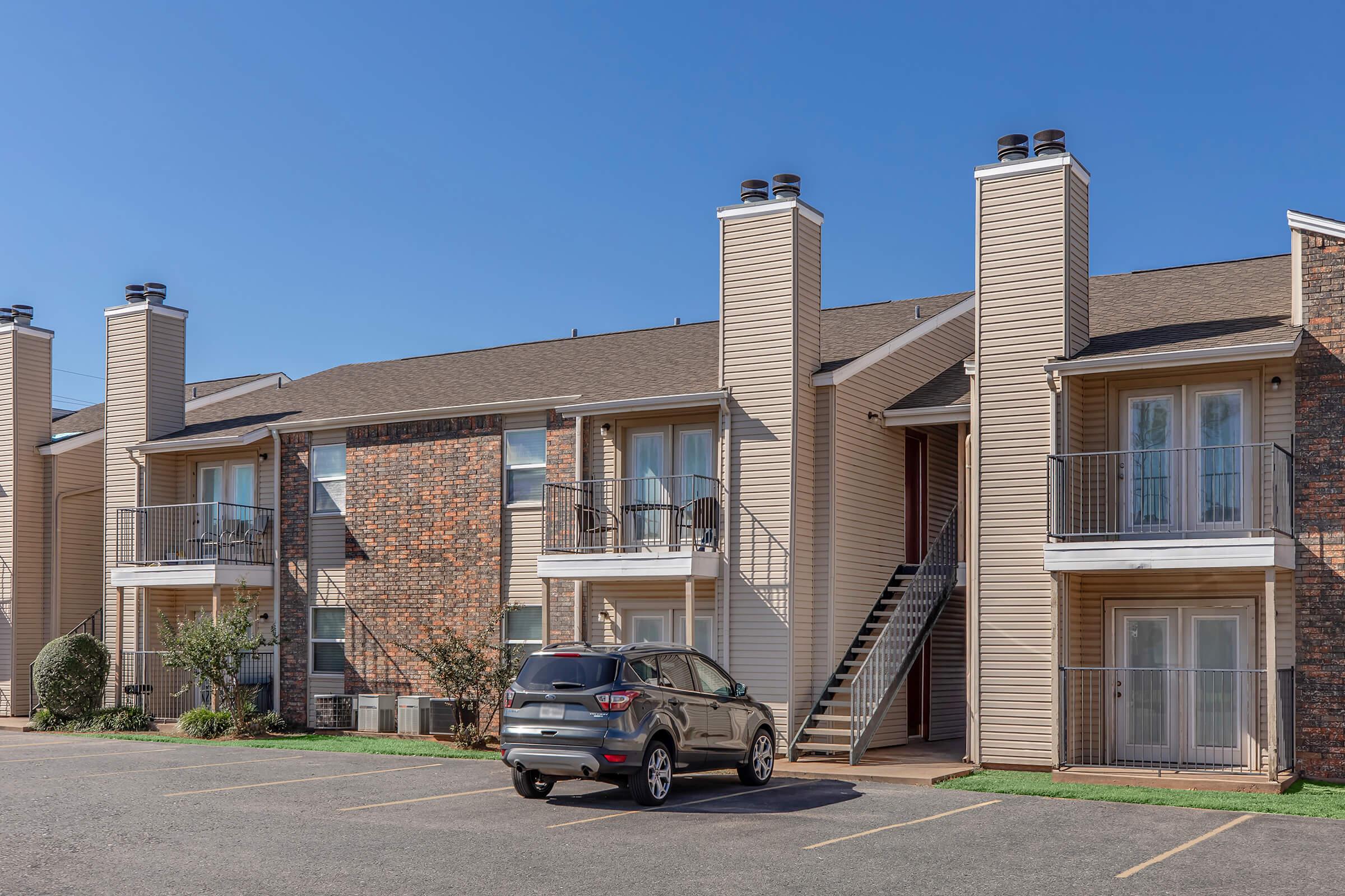 Two-story apartment complex with a mix of brick and siding exteriors, featuring balconies and staircases. A parked SUV is in the foreground, and blue sky with no clouds is visible above. The scene is set in a residential area with a neatly maintained landscape.