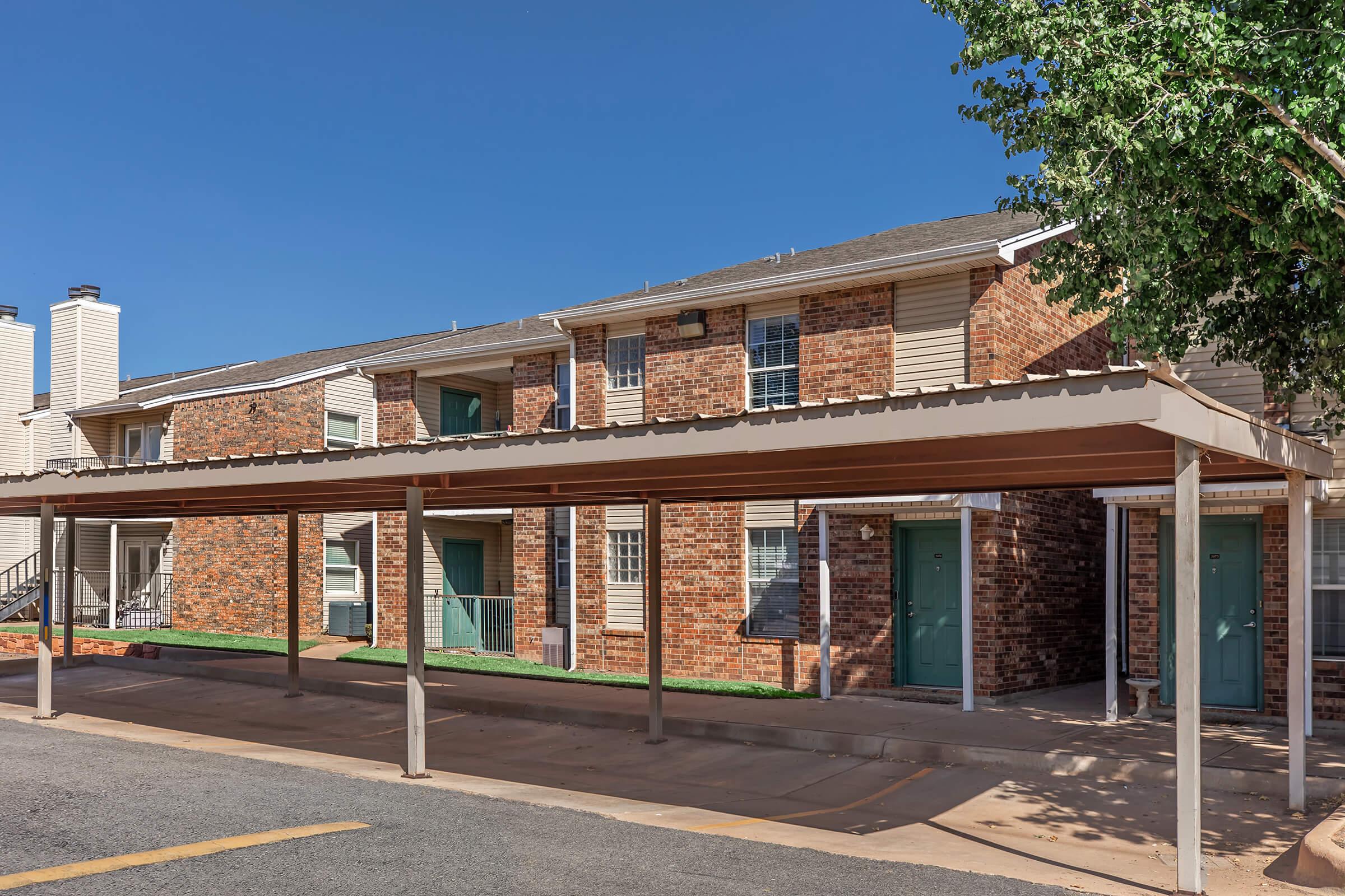 A view of a two-story brick apartment building with green doors, set against a clear blue sky. In the foreground, a covered parking area provides shade, and the landscape features minimal greenery. The exterior showcases a modern yet simple architectural style.