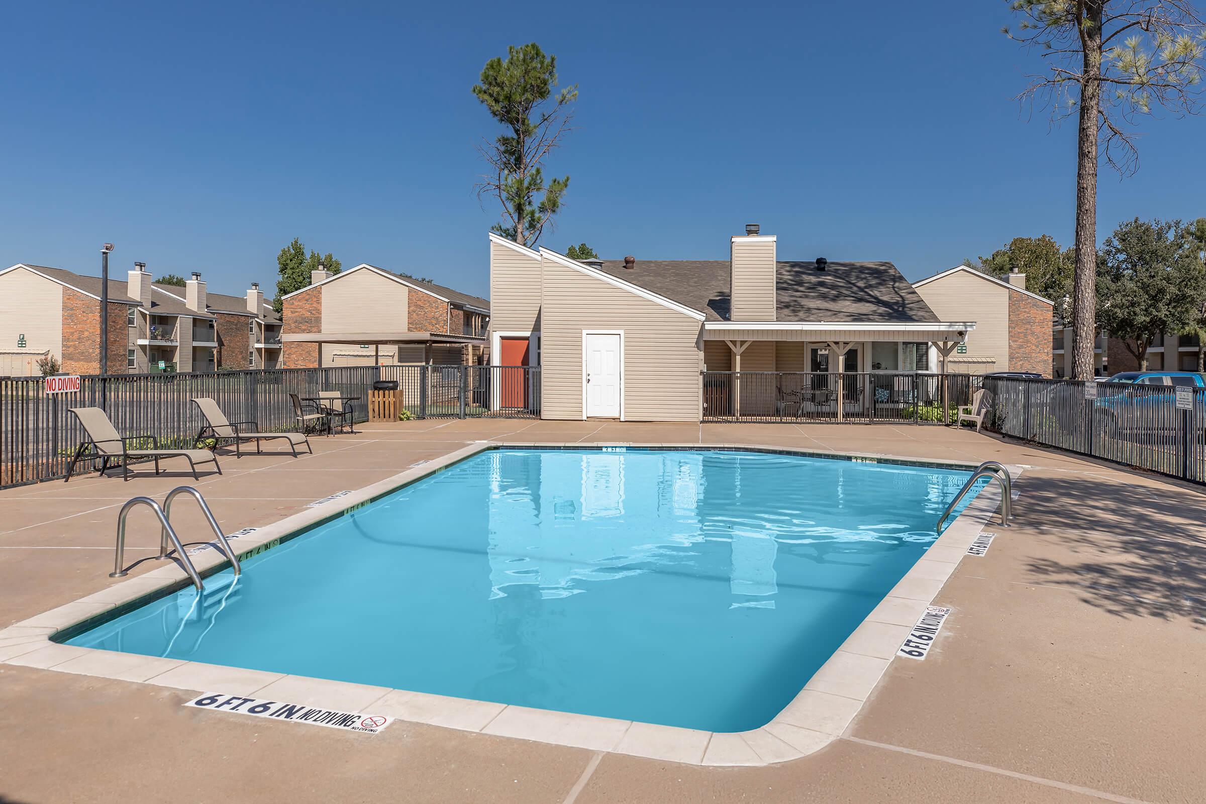 A swimming pool surrounded by a concrete area with lounge chairs. In the background, there are residential buildings and trees. The pool water is clear, and a small building is situated nearby, likely serving as a community area or pool house. The sky is clear and blue.